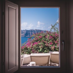 Masterclass about images with an image showing a stunning view out of a window across patio furniture to trees with flowers on it, blue sea, mountains and blue sky