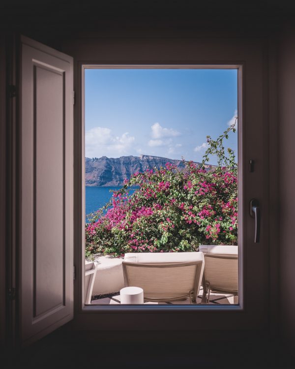 Masterclass about images with an image showing a stunning view out of a window across patio furniture to trees with flowers on it, blue sea, mountains and blue sky