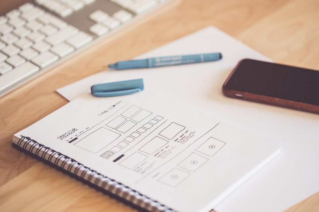 a notepad with words written on a page, with a pen and mobile phone on top of it and keyboard in the background