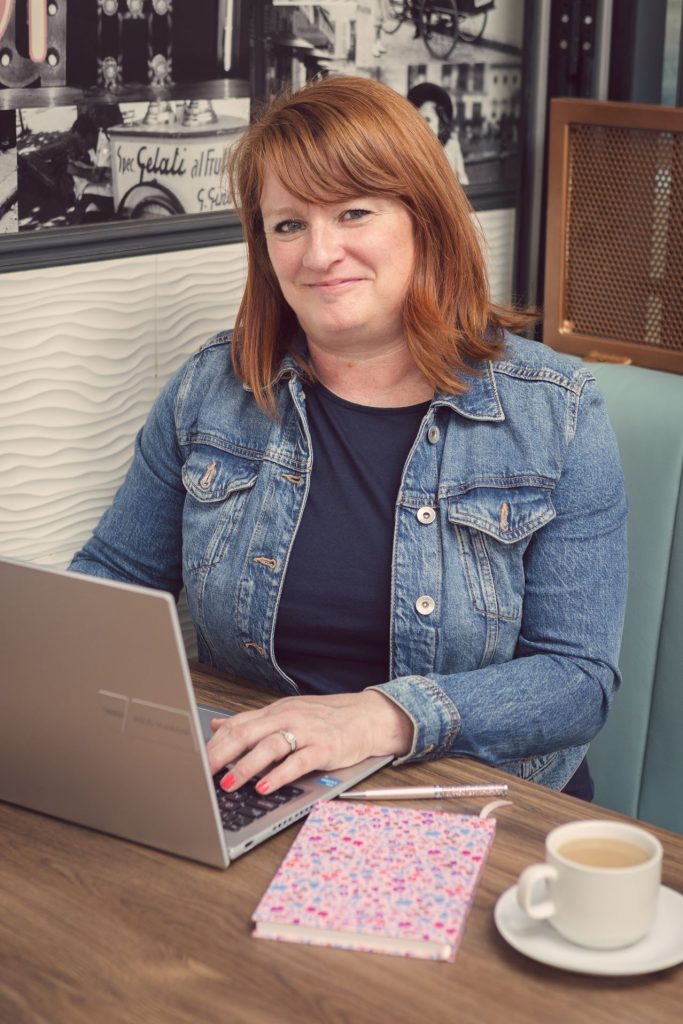 Headshot of Suzy Stanton typing at her laptop with a notebook and cup of tea beside her on a table.