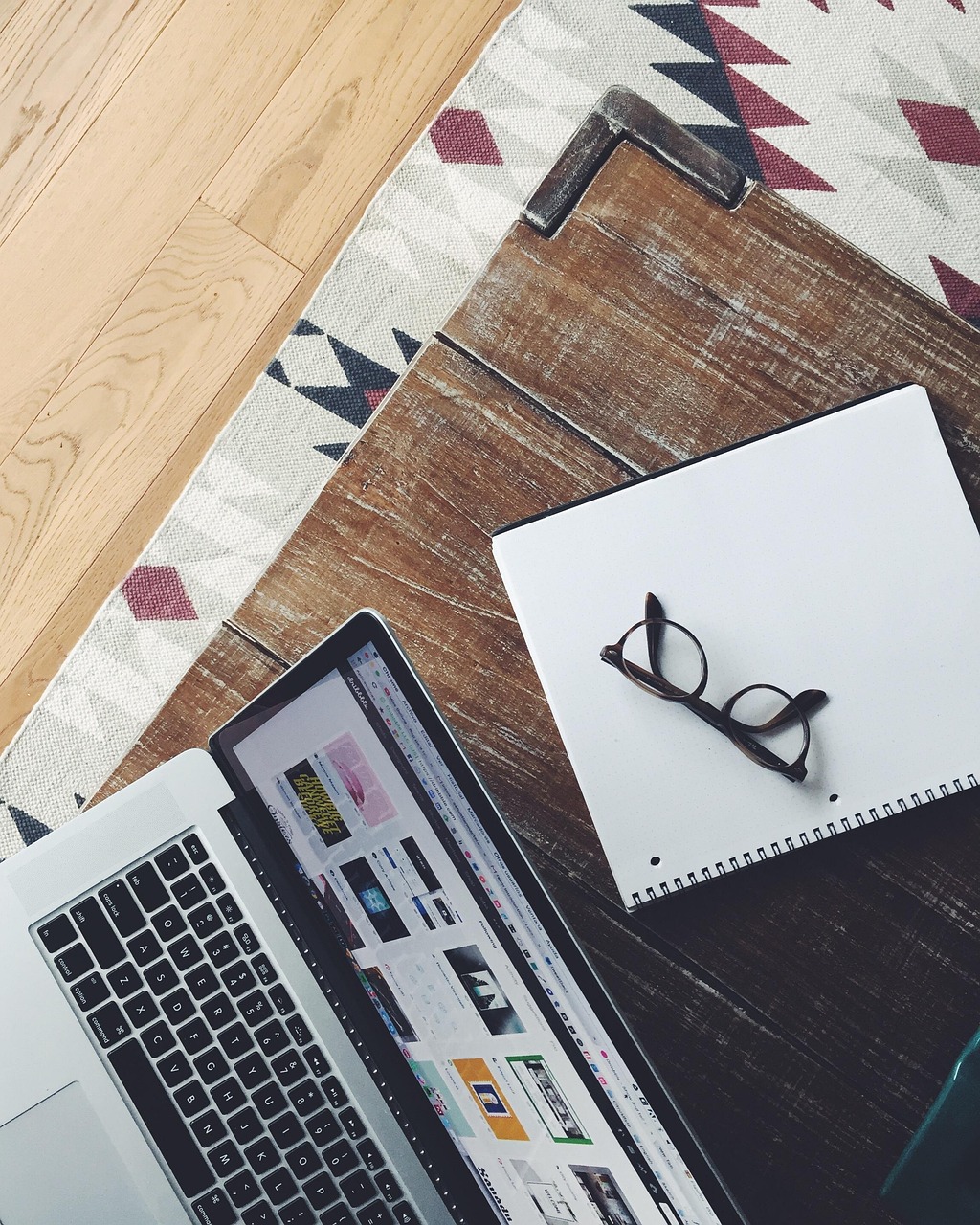 An ariel shot of an open laptop, notebook with a pair of glasses on top, on a dark wooden table
