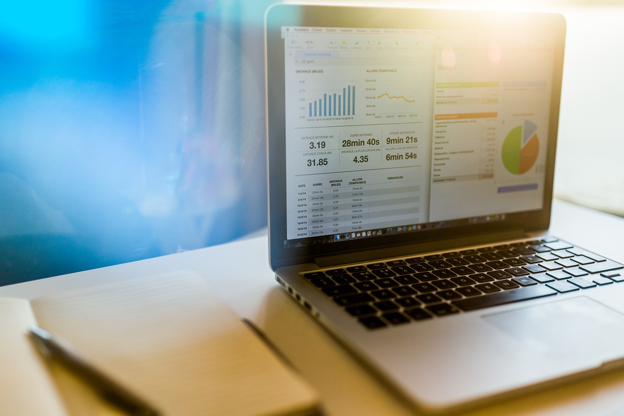 An open laptop with images of statistics on the screen, and an open notebook and pen beside it
