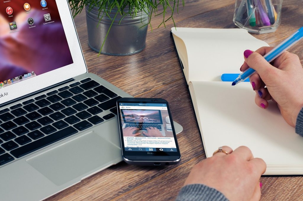 An open laptop with a mobile phone resting on the keyboard and a hand holding a pen writing in a notebook, representing someone creating a visibility strategy 