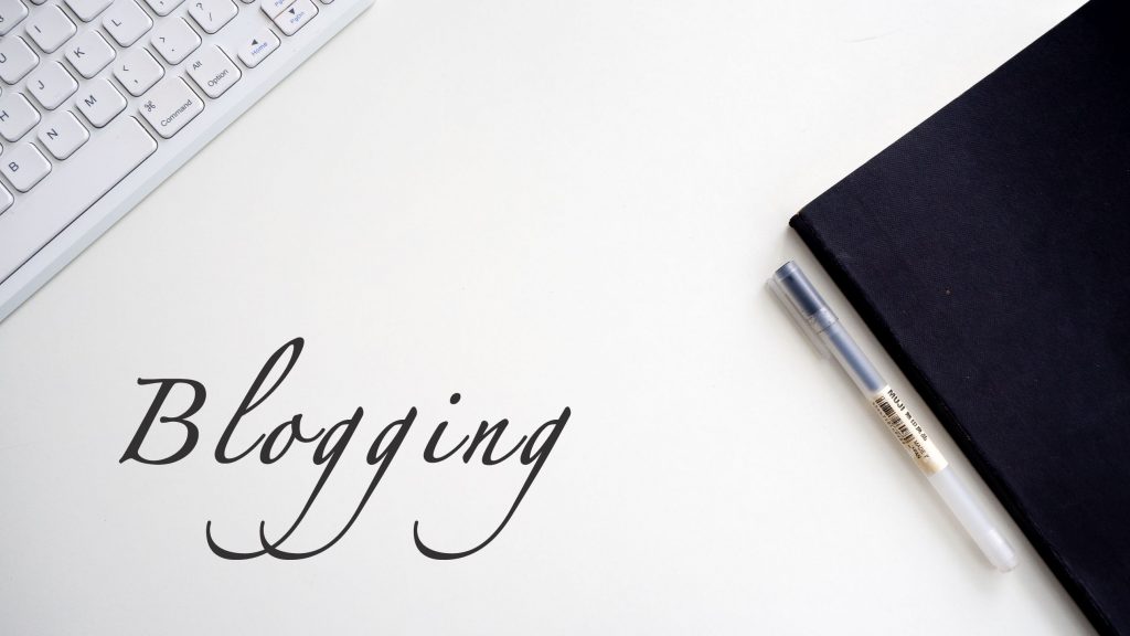 The word blogging written in Calligraphy-style writing on a white background next to part of a keyboard, notepad and pen