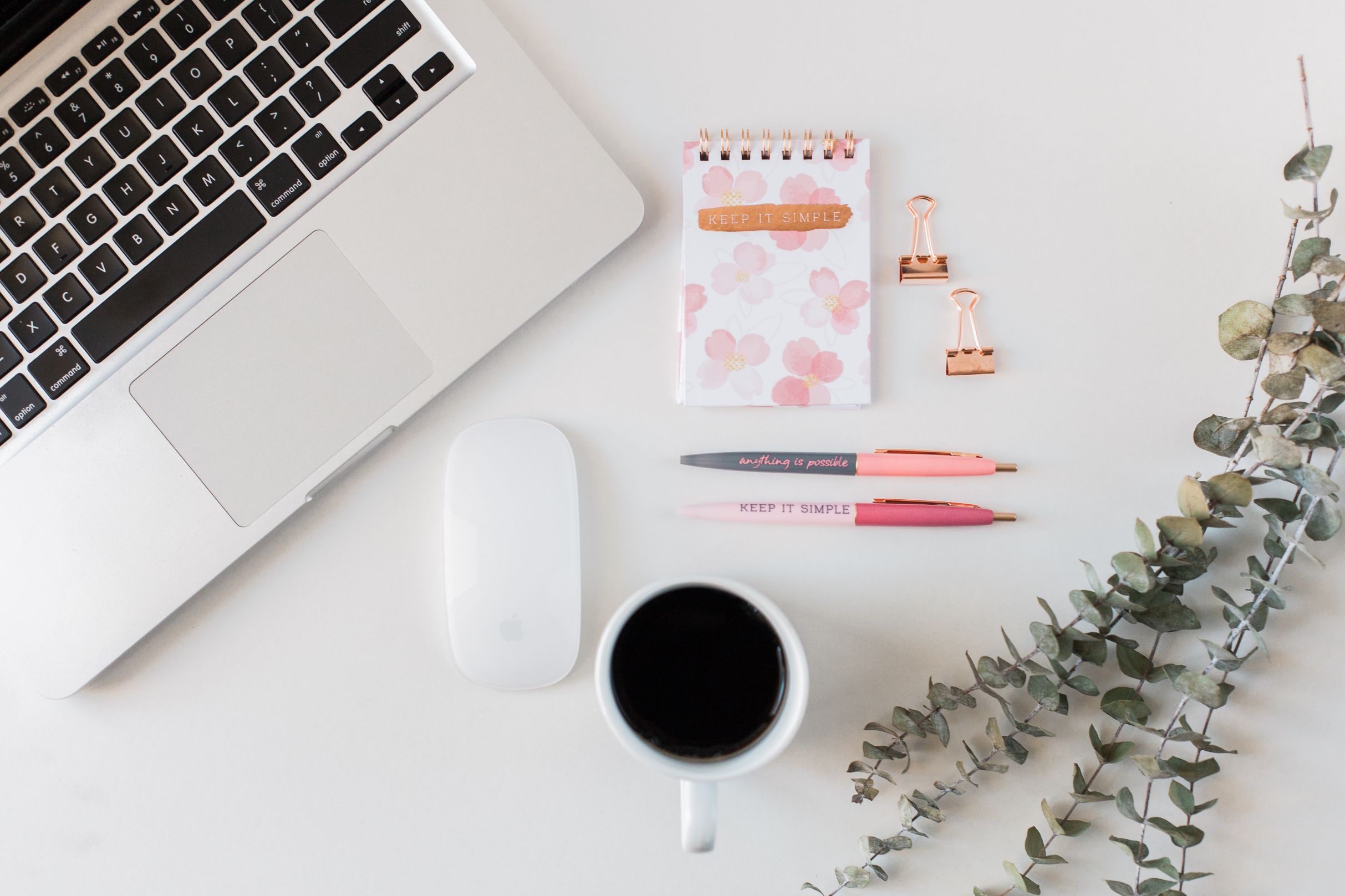 An ariel view of a desk with a keyboard, notepads, pens, coffee in a cup, mouse and plant showing the essentials to get ready to start blogging for business