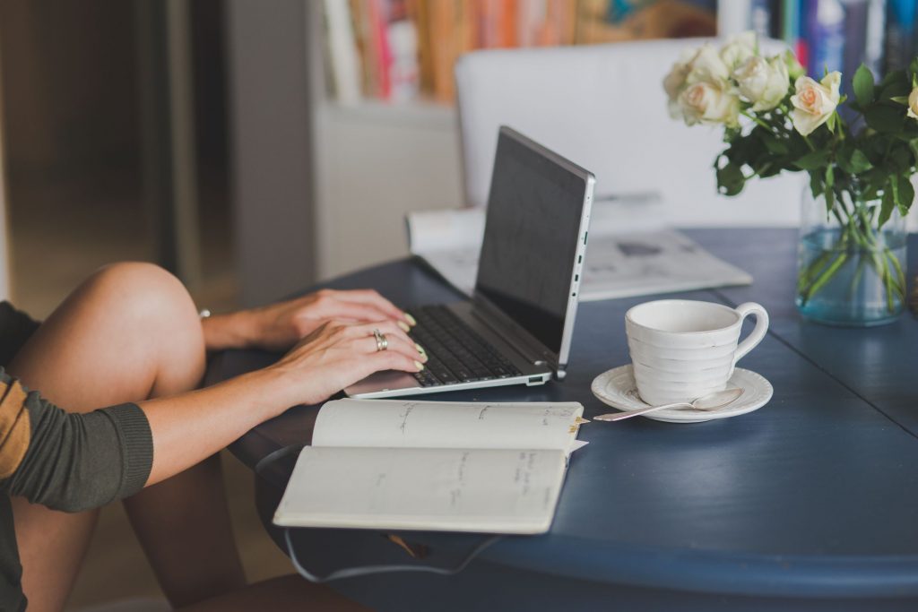 A women typing on an open laptop, with an open notepad beside it, coffee cup and flowers in the background